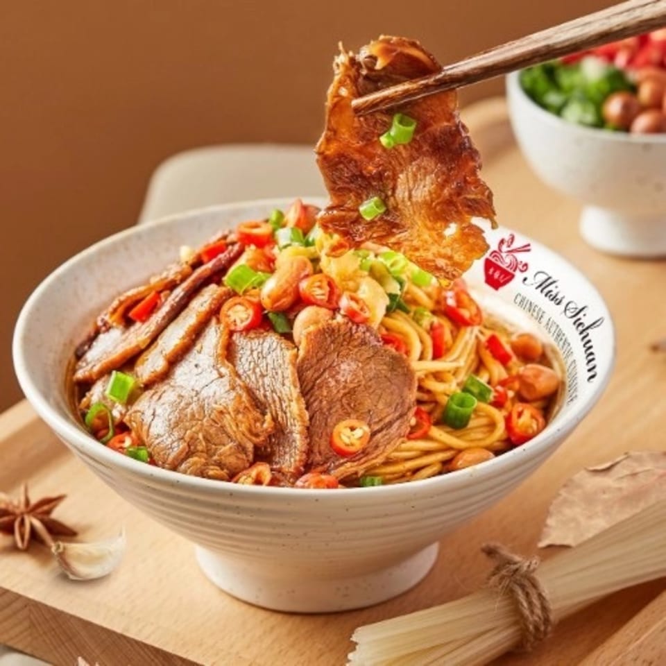Chopsticks lifting a slice of beef from a bowl of Miss Sichuan noodles with chili and peanuts.