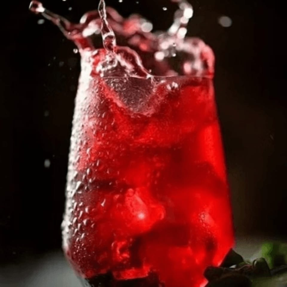 A vibrant red drink with ice splashes out of a glass against a dark background.