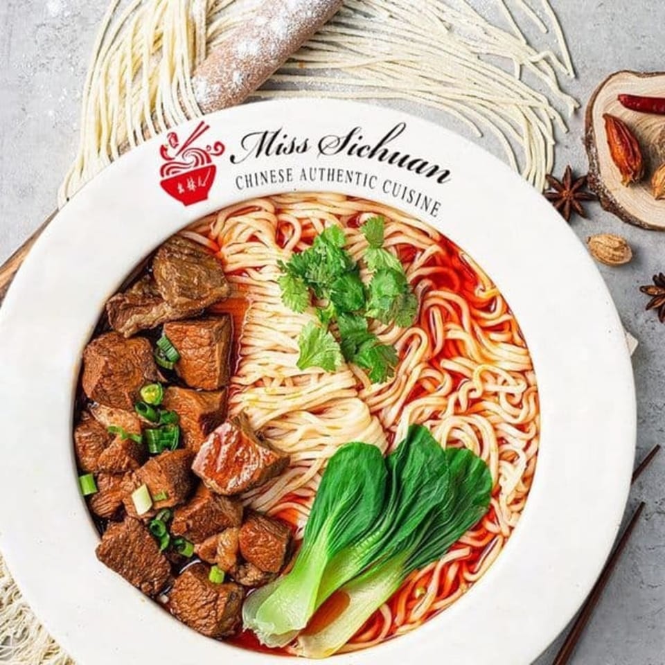 Miss Sichuan beef noodle soup with bok choy and cilantro, surrounded by spices and raw noodles.