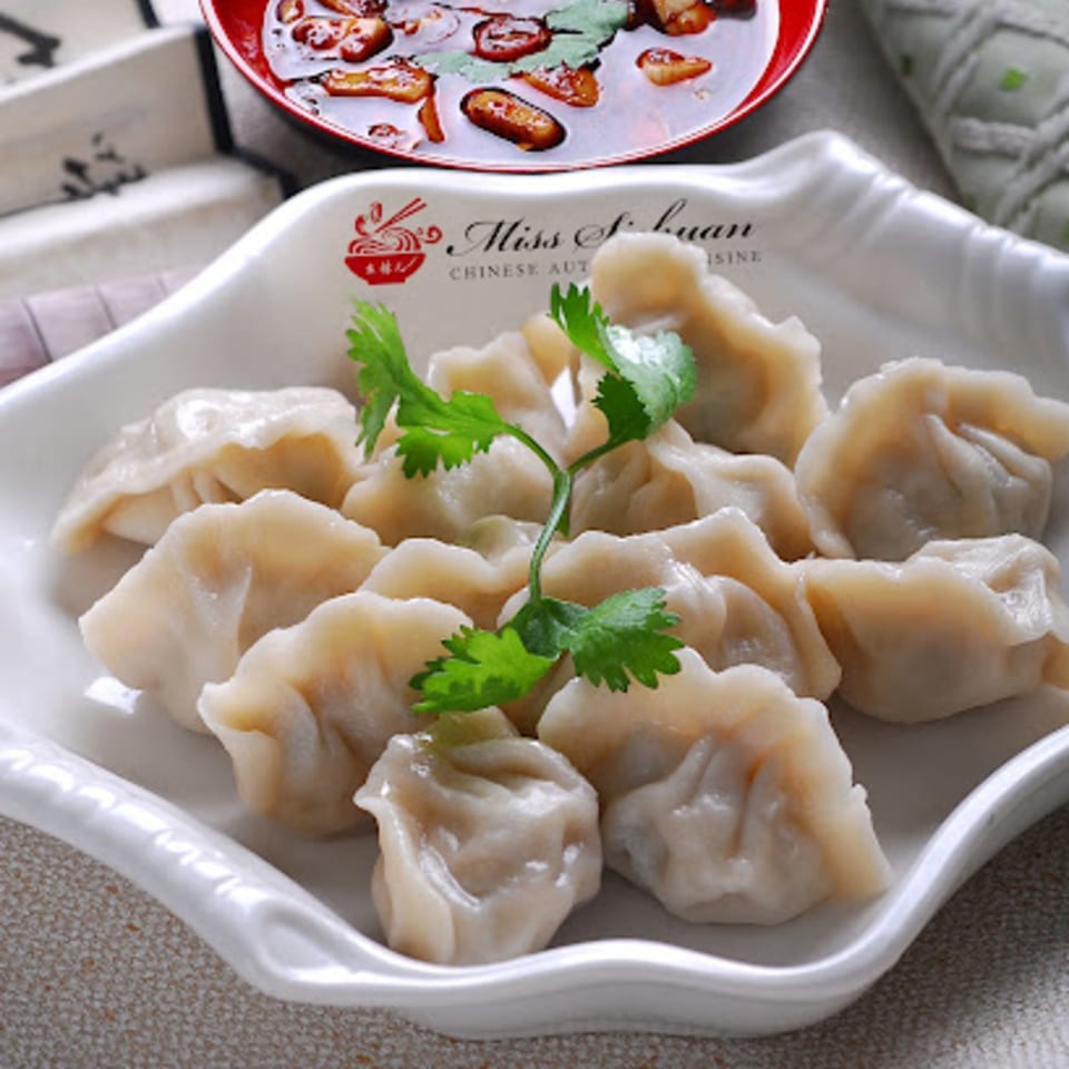 A white Miss Sichuan plate filled with dumplings, garnished with cilantro, and a red bowl of chili oil.