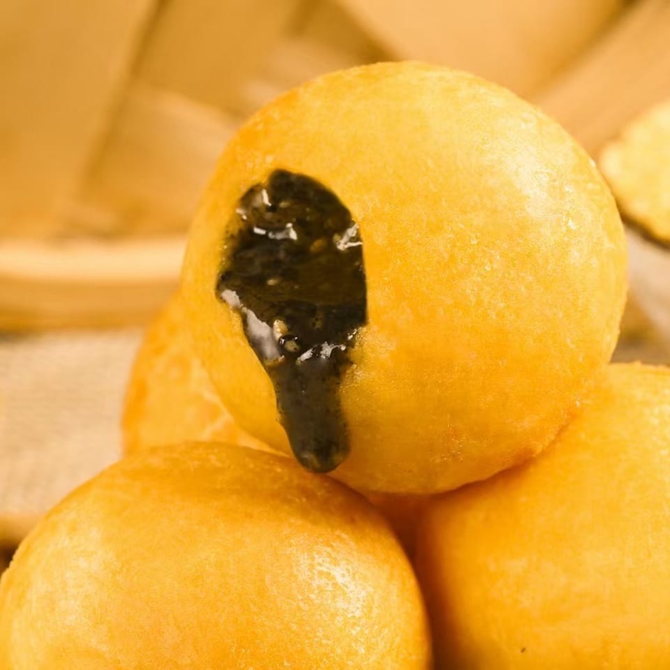 Golden fried sesame balls, one broken open to reveal a dark, gooey filling, on a light background.