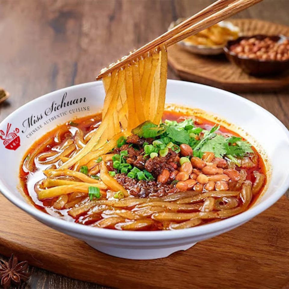 Chopsticks lifting wide noodles from a bowl of Miss Sichuan's spicy Chinese noodles with peanuts and cilantro.